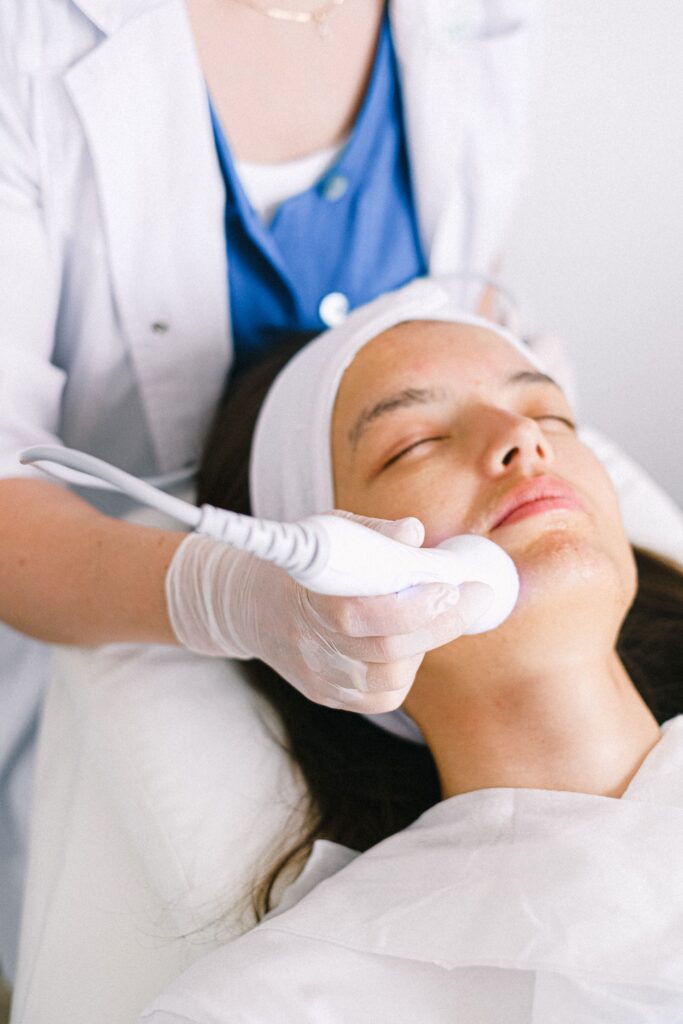 Professional female cosmetologist wearing medical uniform doing ultrasonic face cleaning for relaxed young female customer in modern beauty clinic